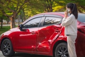 Woman covering face beside damaged car after traffic collision on roadway